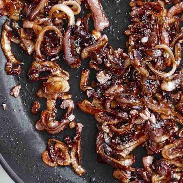 The image shows caramelized shallots in a pan. The shallots have been cooked down until they've reached a deep amber color, indicative of the natural sugars being released and caramelized through a slow and careful cooking process. The edges of the shallots are slightly crisped, suggesting a bit of texture contrast to the soft, jammy centers. The glistening sheen on the shallots is typical of the oil or butter used in the caramelization process, which also adds richness to the flavor. Caramelized shallots are a flavorful addition to many dishes, offering a sweet yet savory depth that enhances the overall taste experience. The pan's dark surface provides a stark contrast to the rich, browned shallots, highlighting their transformation from raw, pungent bulbs to sweet, savory morsels.