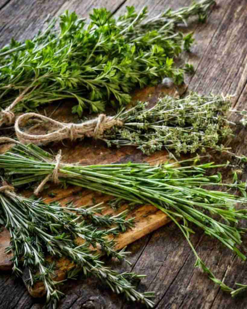 The image shows a collection of fresh herbs on a wooden board. From top to bottom, there appears to be a bundle of flat-leaf parsley, known for its bright green and leafy sprigs, a bunch of thyme with small, delicate leaves, and lastly, a bundle of oregano, which can be identified by its slightly rounder leaves compared to thyme. Each herb is tied with a piece of twine, indicating they have been freshly picked and are possibly prepared for drying or immediate use in cooking. The wooden background provides a rustic and organic backdrop, highlighting the herbs' natural state. Perfect for our infused cream!