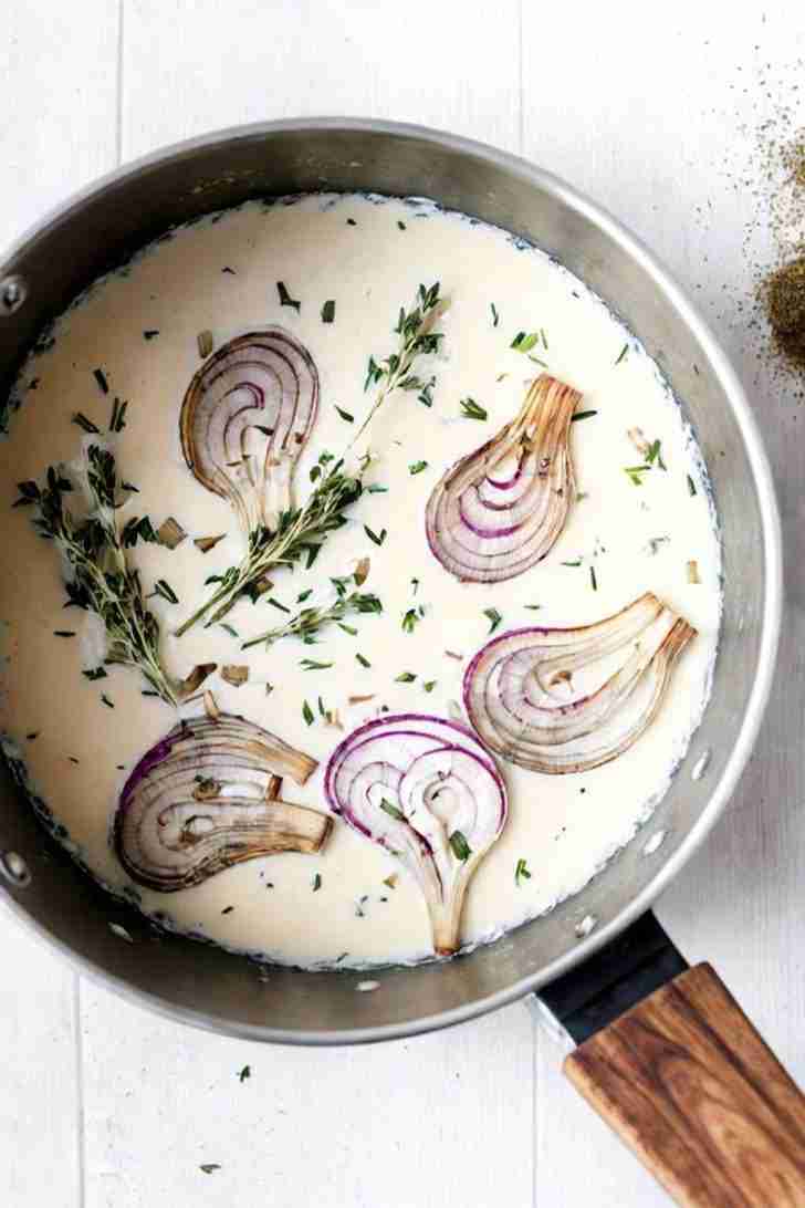 The photo displays a pot filled with infused cream. Slices of red onion are partially submerged in the cream, and fresh thyme sprigs are visible throughout the mixture. The combination of onions and thyme suggests a savory profile for the infusion. The pot has a wooden handle and sits against a light, neutral background which accentuates the contents.