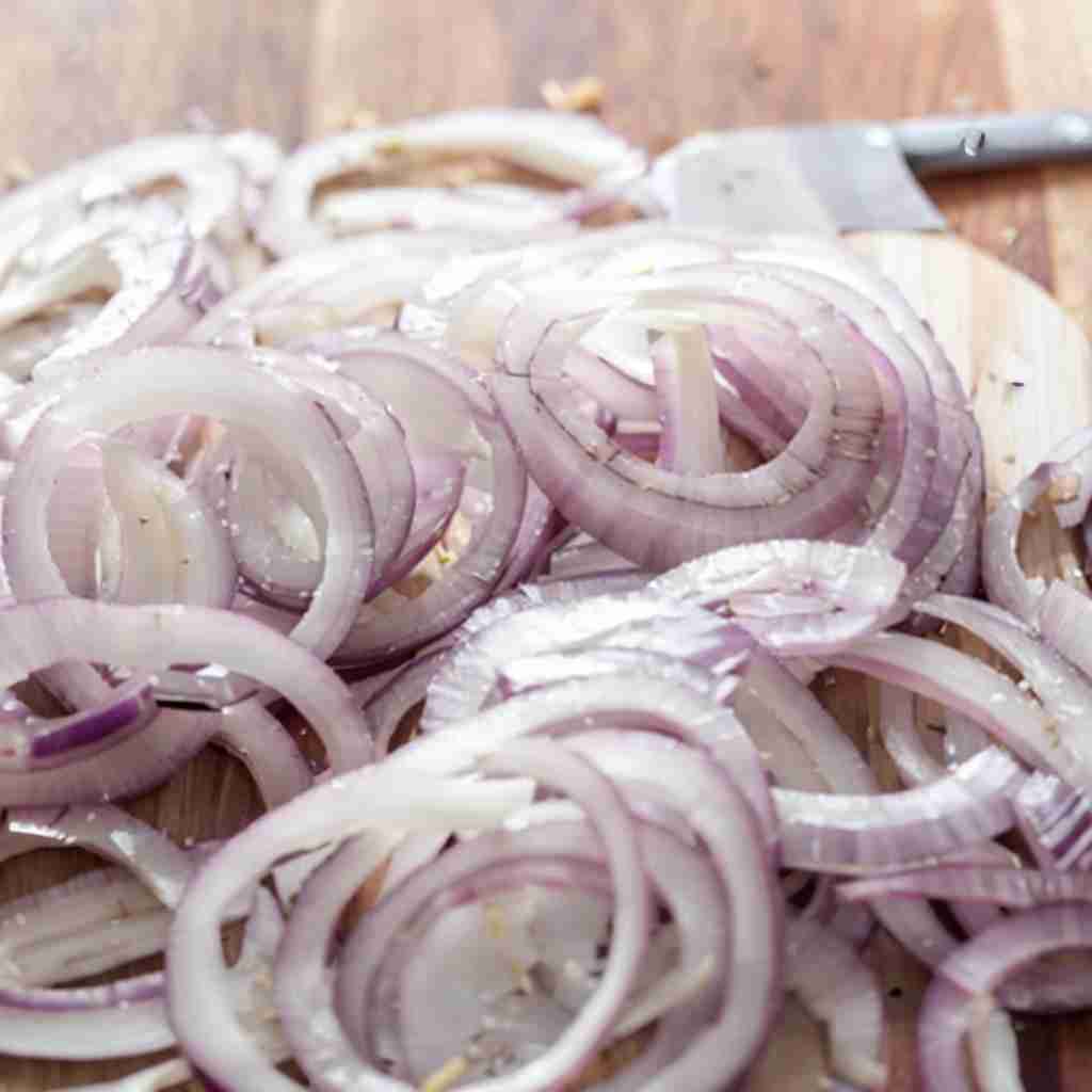 thinly sliced shallots arranged on a wooden chopping board. The shallots are fresh and raw, with their characteristic purple and white hues. They are cut into rings, displaying the typical layered structure of alliums. There's a chef's knife visible in the background, suggesting that the slicing has just been done. The shallots appear ready for cooking, ready to become caramelized shallots, as their thin slicing would allow them to cook quickly and evenly. The wooden board and the knife hint at an ongoing preparation in a kitchen setting. The focus and brightness of the image highlight the freshness of the shallots, which are a staple ingredient in many culinary creations for their mild yet distinct flavor.