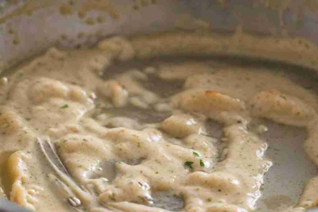 In this close-up image, we observe a roux-based sauce in the midst of cooking. The sauce has a creamy beige color, indicative of the flour and fat cooked together until they’ve reached a light, nutty hue. Scattered specks of herbs, perhaps finely chopped parsley, pepper the sauce, hinting at the infusion of flavor. The texture appears luxuriously smooth and thick, suggesting the roux has been expertly whisked into a liquid, possibly stock or milk, and brought to the right consistency without any visible lumps. There's a glistening sheen on the surface, reflecting meticulous cooking which has allowed the starch granules to swell and burst, creating this velvety consistency. The edges of the sauce exhibit a slightly foamy quality, perhaps from a gentle simmer, which is key to developing depth in flavor and achieving full thickening power. This sauce could be the foundation of a classic dish, like a gratin, béchamel for a lasagna, or a base for a luxurious chicken or seafood entrée. The mastery of roux showcased here is fundamental to culinary arts, combining the science of cooking with the craft of sauce-making.