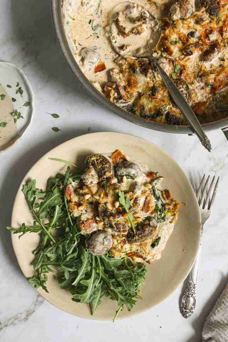 Plate of cheesy mushroom and bacon gratin served with arugula, garnished with toasted pine nuts, next to a skillet of the same dish.