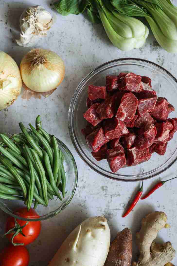 A variety of fresh ingredients laid out for beef sinigang, including cubed raw beef, onions, green beans, bok choy, garlic, tomatoes, daikon radish, ginger, and red chilies.