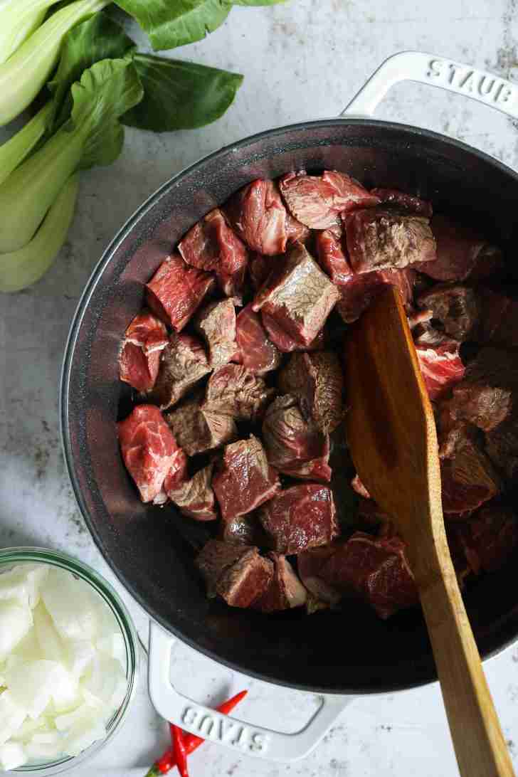 Chunks of beef browning in a pot, stirred with a wooden spoon, with bok choy and a bowl of diced onions nearby, ready to be added.