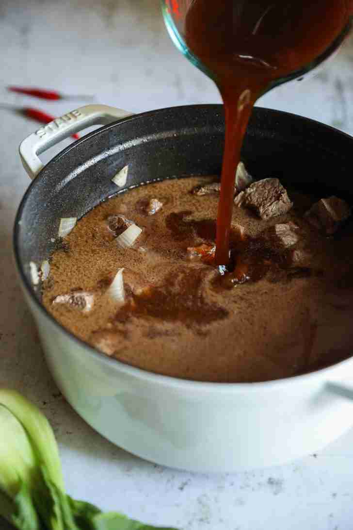 Rich brown broth being poured into a pot filled with seared beef chunks and onions, with bok choy and red chilies in the background.