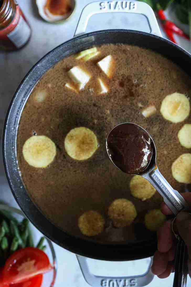 A spoonful of tamarind paste being added to a pot of simmering broth with chunks of taro floating in the soup, surrounded by green beans and tomatoes.