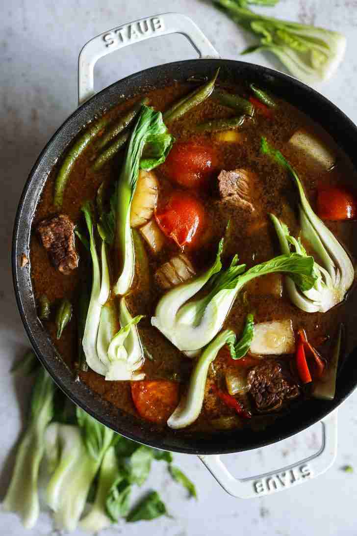 A pot of beef sinigang simmering with vibrant bok choy, green beans, tomatoes, and tender chunks of beef in a rich broth.