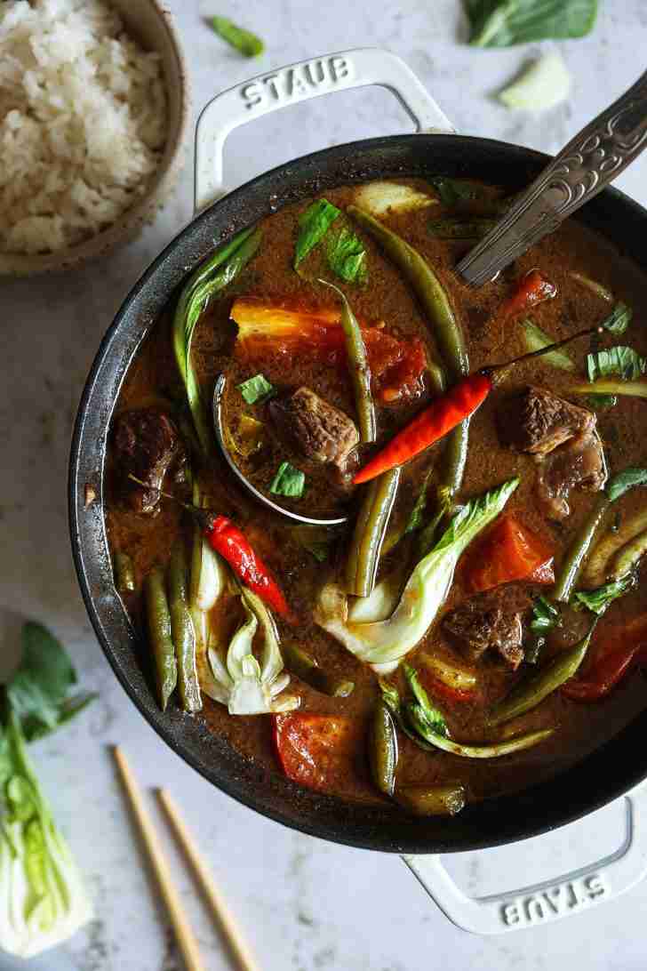 A finished pot of beef sinigang filled with green beans, bok choy, red chilies, tomatoes, and tender beef, served alongside a bowl of steamed white rice.