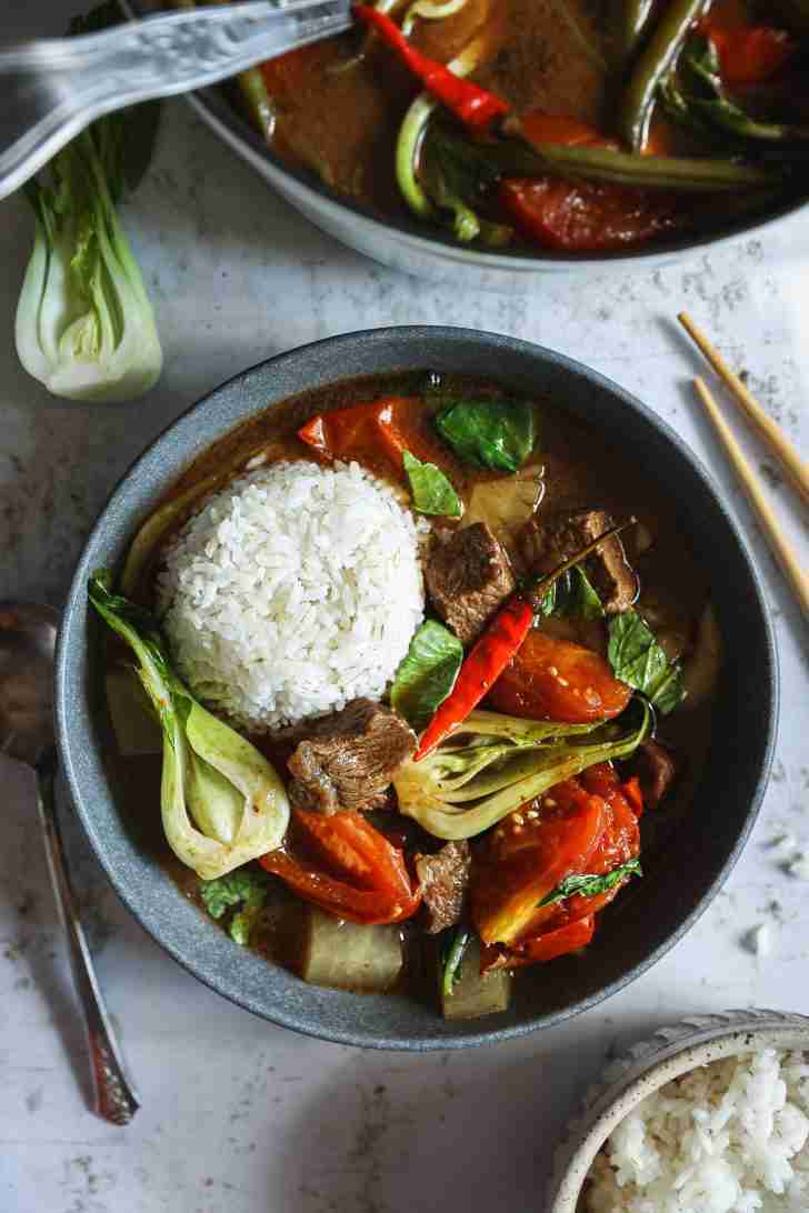 A bowl of beef sinigang served with a mound of steamed rice, featuring vibrant bok choy, tender beef chunks, tomatoes, and a red chili pepper.