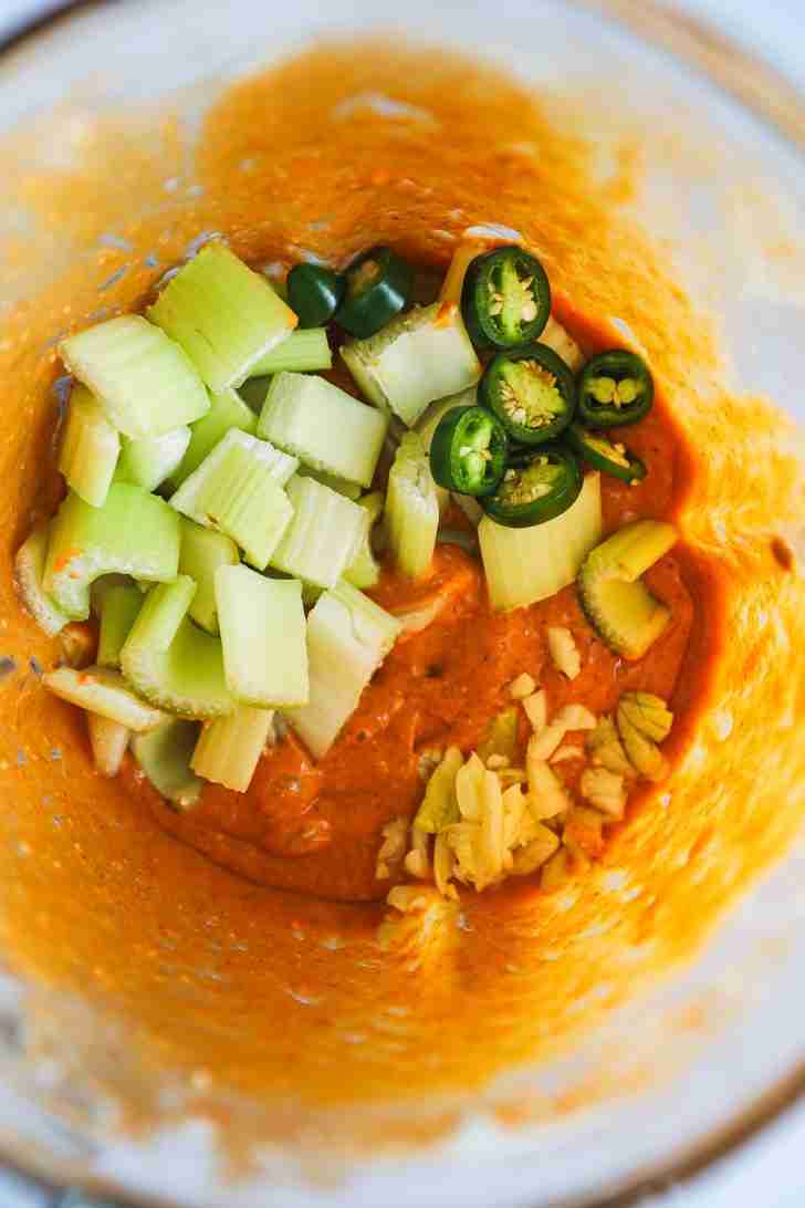 Celery, sliced pepper, and minced garlic added to the creamy orange Blue Heat Wing Sauce mixture inside a blender, ready to be pulsed.