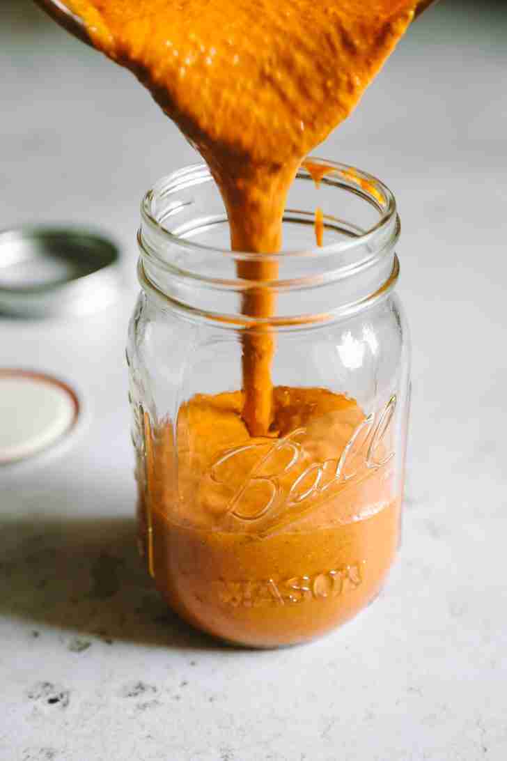 The finished Blue Heat Wing Sauce being poured into a glass mason jar for storage, showcasing its smooth and vibrant orange texture.