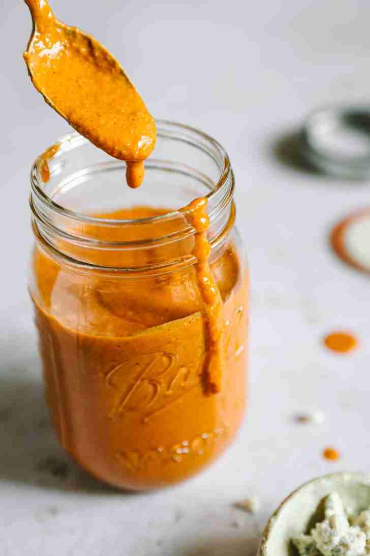 A spoon drizzles the creamy Blue Heat Wing Sauce into a mason jar, with some of the vibrant orange sauce spilling over the jar’s edge.