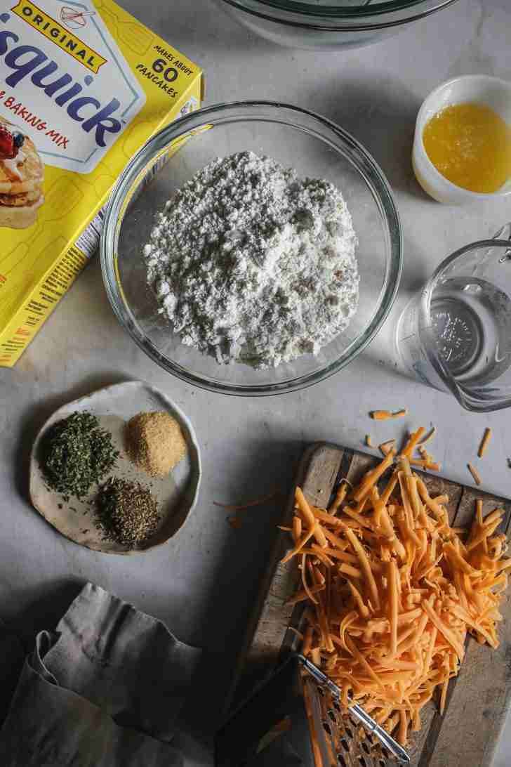 Ingredients for Classic Cheese Biscuits arranged on a counter: Bisquick mix in a glass bowl, grated cheddar cheese, seasonings, melted butter, and a measuring cup of water.