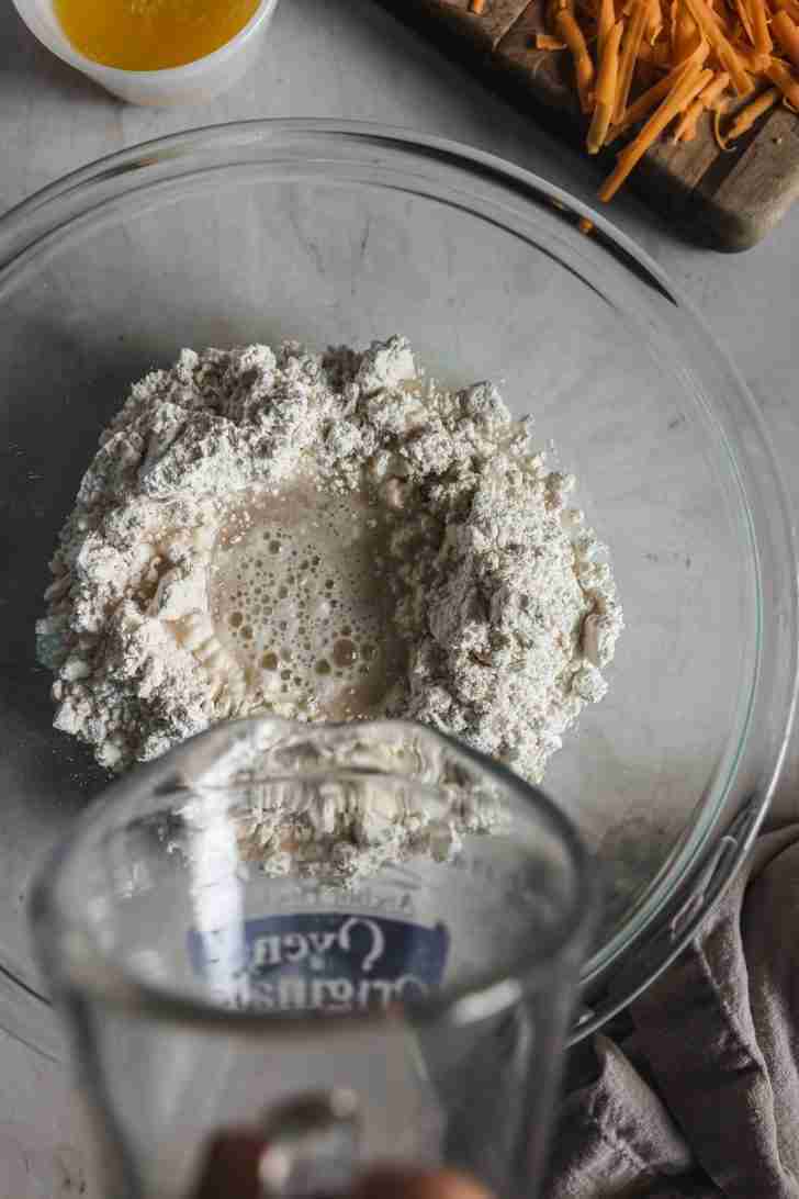 Water being poured into a glass bowl filled with Bisquick mix to form the biscuit dough, with melted butter and grated cheddar cheese set aside.