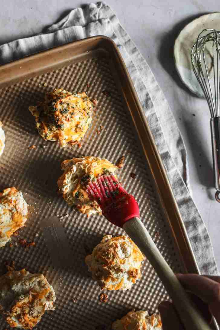 Freshly baked cheese biscuits on a baking sheet being brushed with a garlic herb butter mixture.