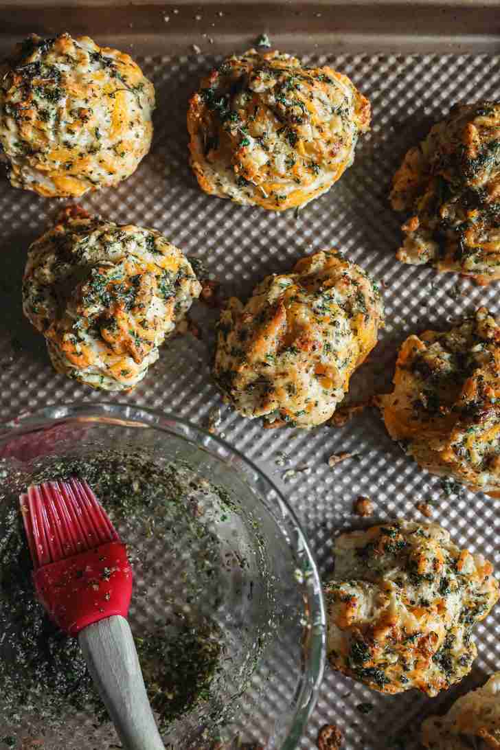 Golden biscuits topped with melted cheese and herbs are arranged on a baking sheet, with a bowl of leftover herb butter and a red brush nearby.