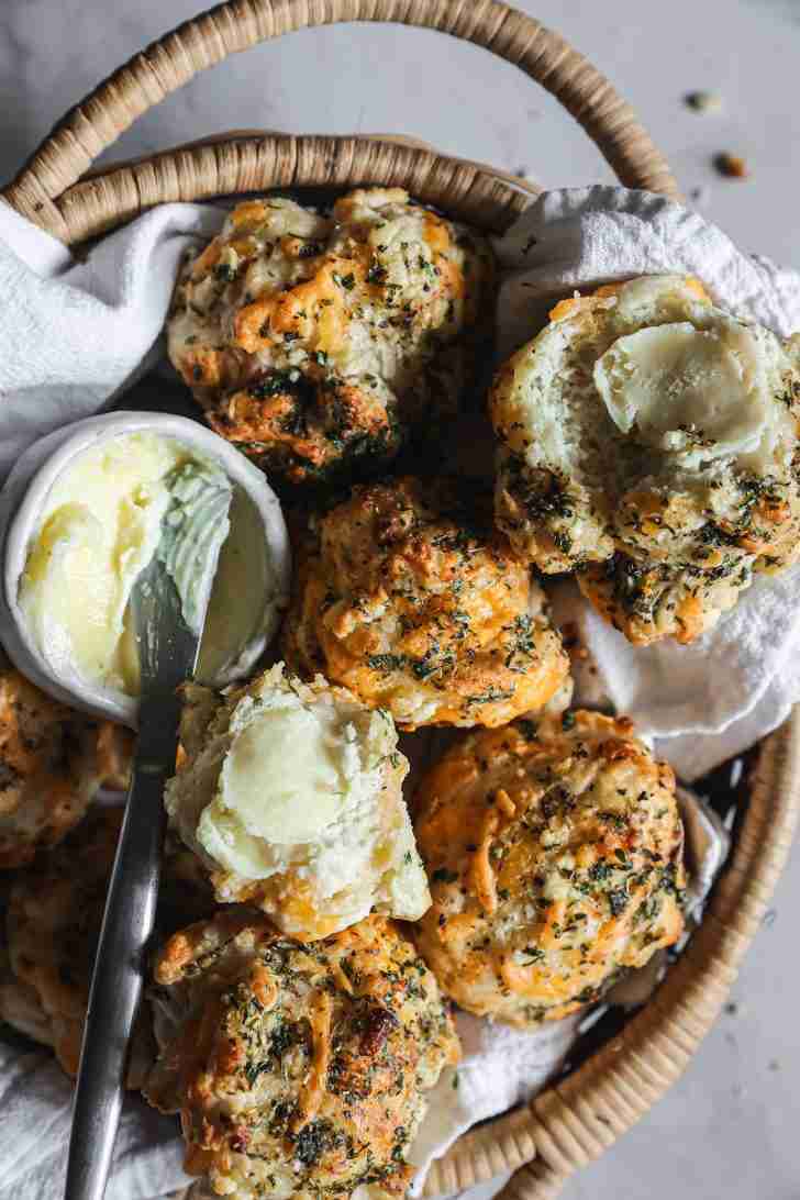 A basket filled with warm cheese biscuits, some halved and topped with butter, alongside a small dish of spreadable butter and a butter knife.