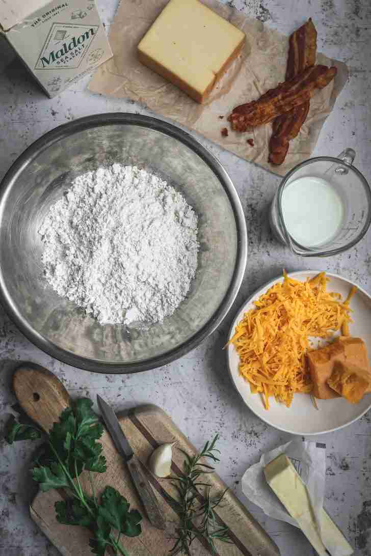 A bowl of Bisquick mix sits among cheddar, Gruyère, bacon, herbs, garlic, butter, and buttermilk, ready to make savory cheese biscuits.