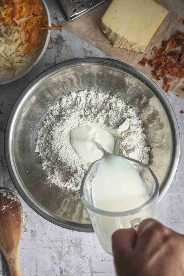 Buttermilk is poured into a bowl of Bisquick mix, with shredded cheddar, Gruyère, bacon crumbles, and a wooden spoon nearby, ready to be mixed.