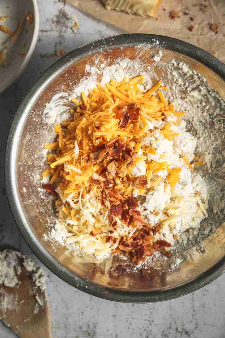 A bowl filled with Bisquick mix is topped with shredded cheddar, Gruyère, and crispy bacon bits, ready to be folded into the dough.