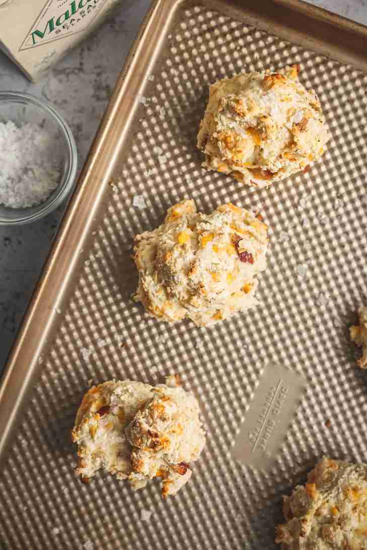 Freshly baked biscuits with a golden, lightly crisp top are on the baking sheet, sprinkled with flaky sea salt for a savory finish.