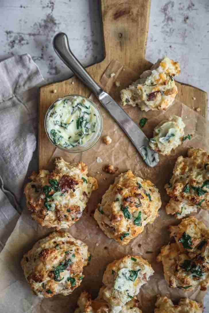 Golden brown cheese biscuits, topped with fresh herbs, are arranged on a wooden board alongside a dish of creamy herb butter ready for spreading.