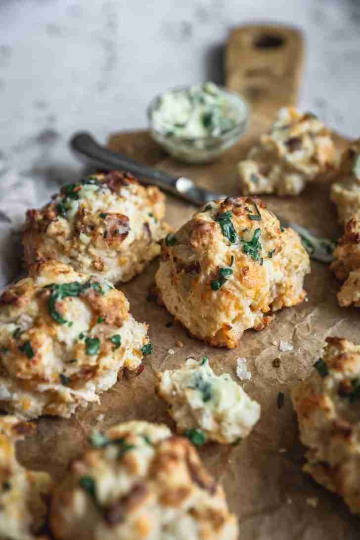 Close-up of golden, flaky biscuits garnished with fresh herbs. One biscuit is broken open, showing its soft interior next to a knife and herb butter.