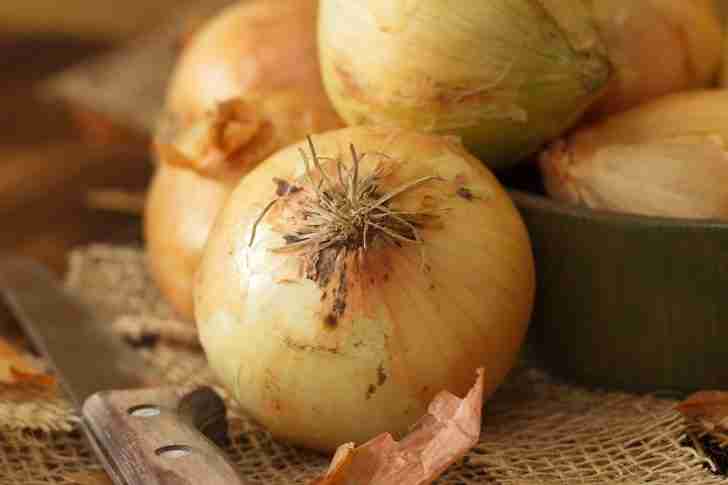 A close-up of whole yellow onions resting on a textured burlap surface, with a wooden-handled knife placed beside them, ready for slicing.