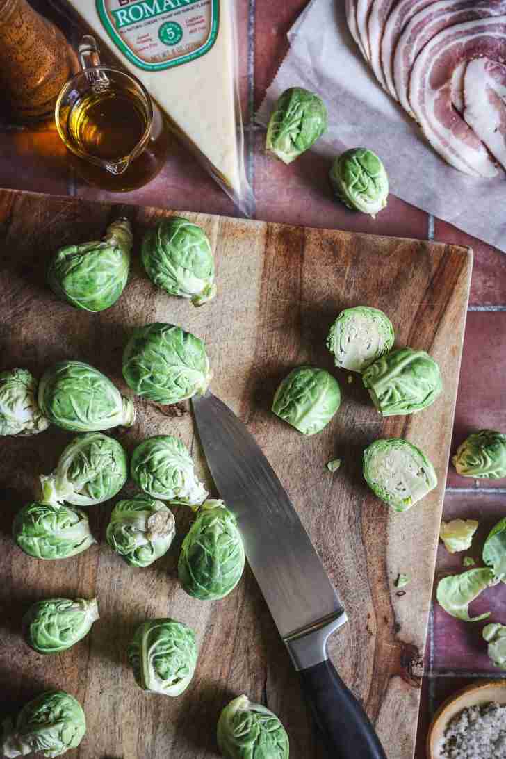 Fresh Brussels sprouts being trimmed and halved on a cutting board with a knife, alongside pancetta and a block of Romano cheese.