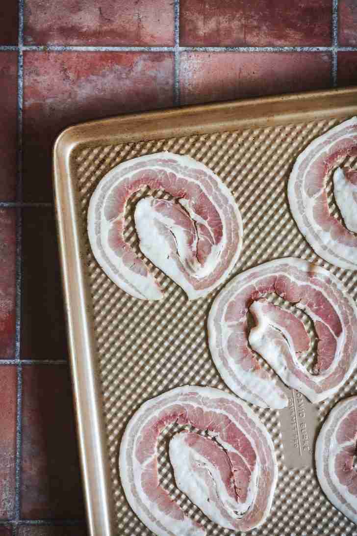 Thinly sliced pancetta arranged in circular patterns on a textured baking sheet, prepared to be baked in the oven until browned and crispy.