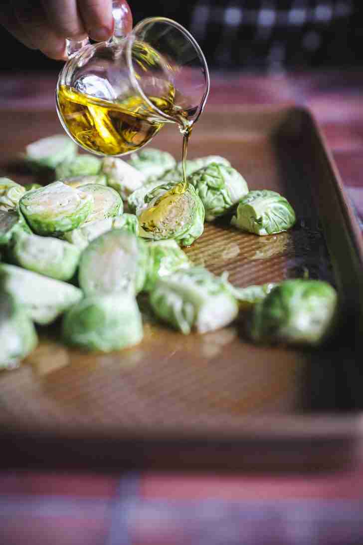 Brussels sprouts halved and spread on a baking sheet as a hand pours golden cooking oil over them, preparing them for roasting in the oven.