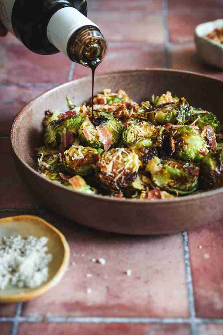 Roasted Brussels sprouts topped with crispy pancetta and grated Romano cheese, being drizzled with balsamic glaze, ready to serve in a bowl.