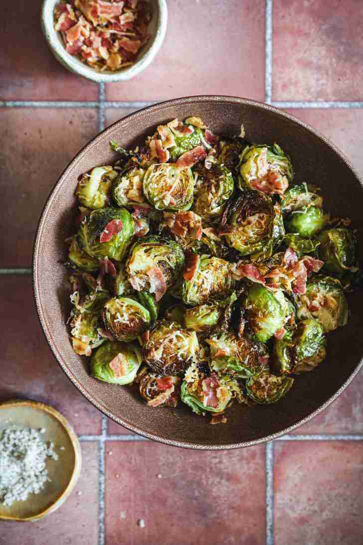 A bowl of roasted Brussels sprouts mixed with crispy pancetta and topped with grated Romano cheese, served with a side of extra pancetta crumbles and salt.