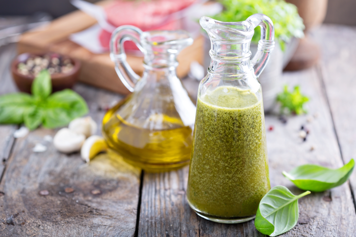 A glass pitcher filled with fresh, homemade salad dressing sits on a rustic wooden table next to a bottle of olive oil, basil leaves, and garlic cloves