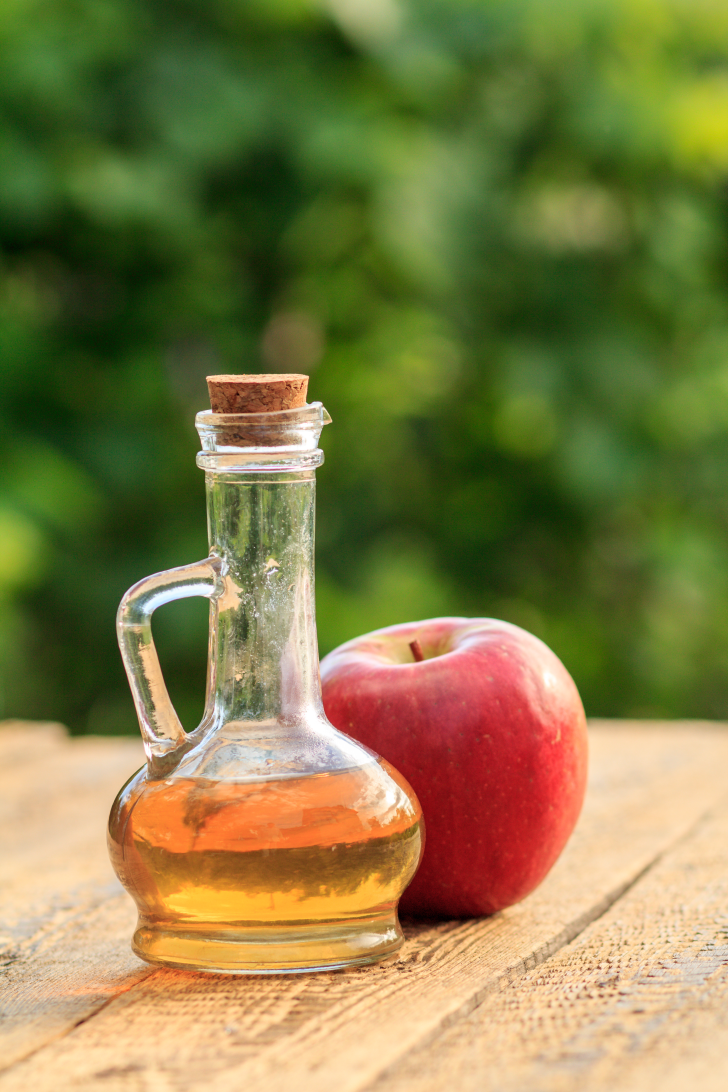 A glass bottle of apple cider vinegar with a cork stopper sits on a wooden table, next to a bright red apple, with a lush green background.