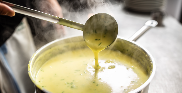 A ladle pours rich, steaming Béarnaise sauce from a pot, revealing its creamy texture and fresh herbs