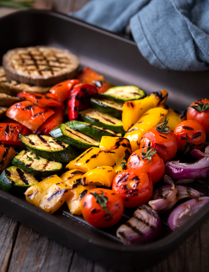 A vibrant assortment of grilled vegetables, including zucchini, bell peppers, cherry tomatoes, and red onions, are beautifully charred and arranged in a roasting pan.