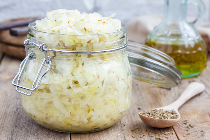 A jar filled with homemade sauerkraut sits on a rustic wooden table, next to a wooden spoon of spices and a bottle of olive oil in the background.