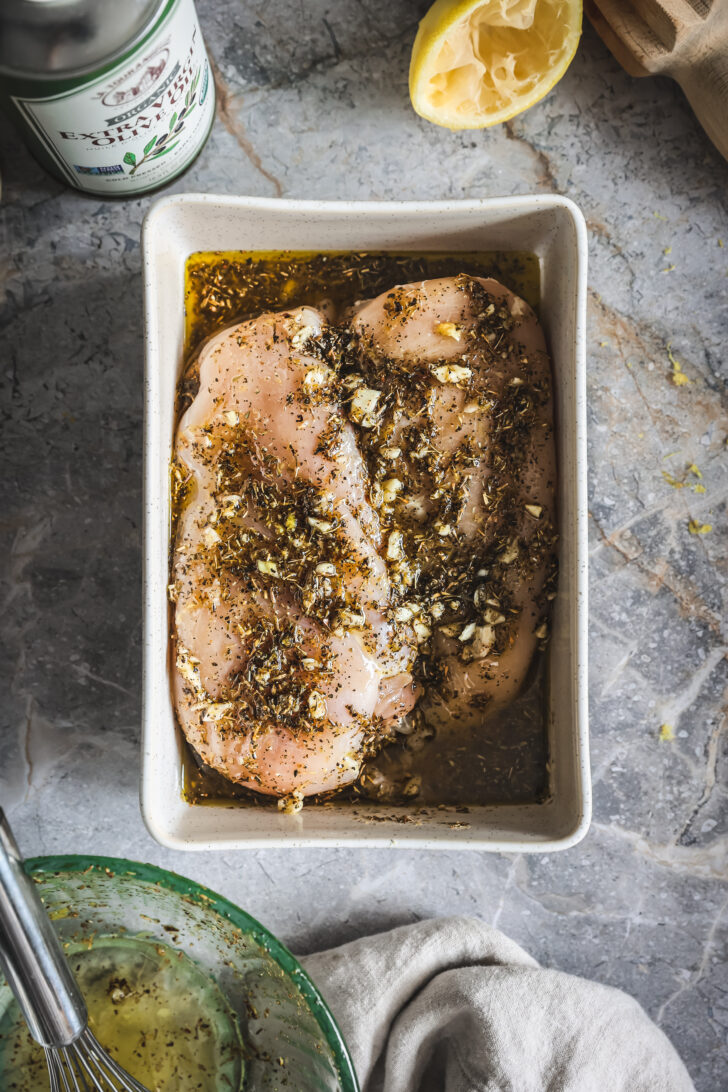 Chicken breasts marinating in lemon herb marinade with minced garlic, olive oil, and spices in a white dish.