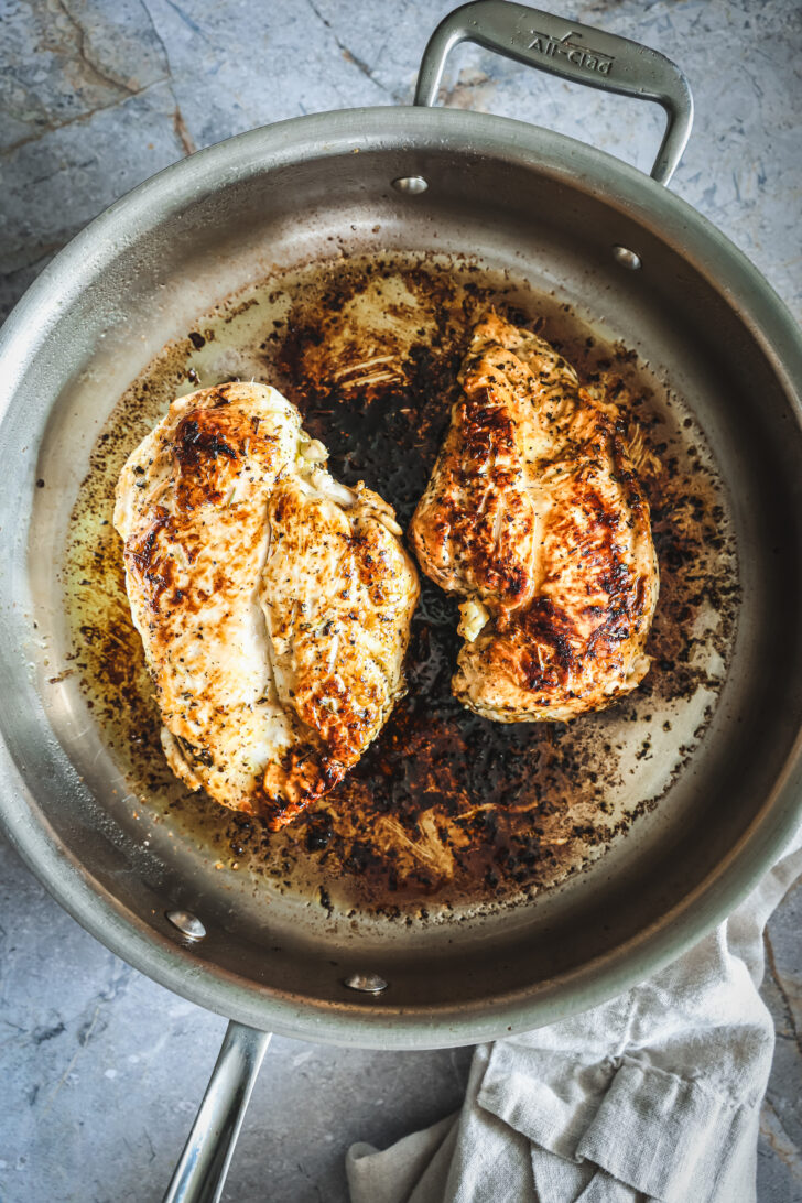 Two golden-brown, seared chicken breasts cooking in a stainless steel skillet with caramelized bits