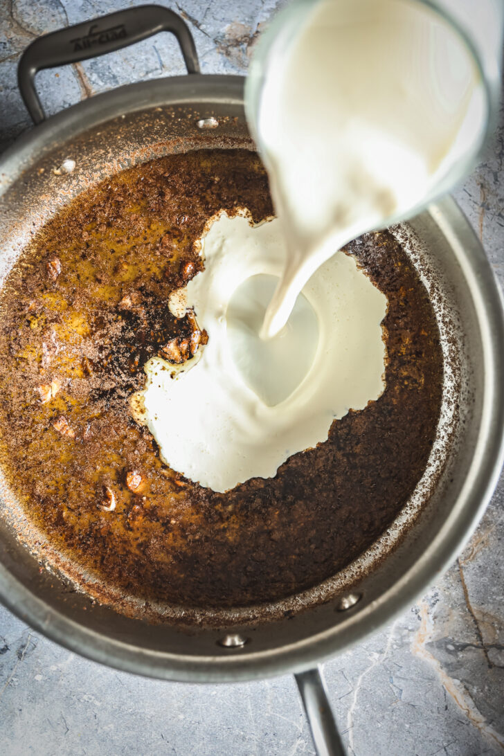 Heavy cream pouring into a skillet with browned pancetta drippings, starting the base for a creamy Alfredo sauce.