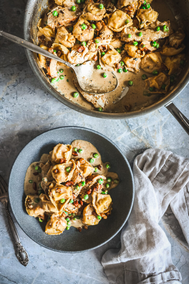 Serving of chicken tortellini with Alfredo sauce, peas, and pancetta in a bowl, with skillet and spoon nearby.