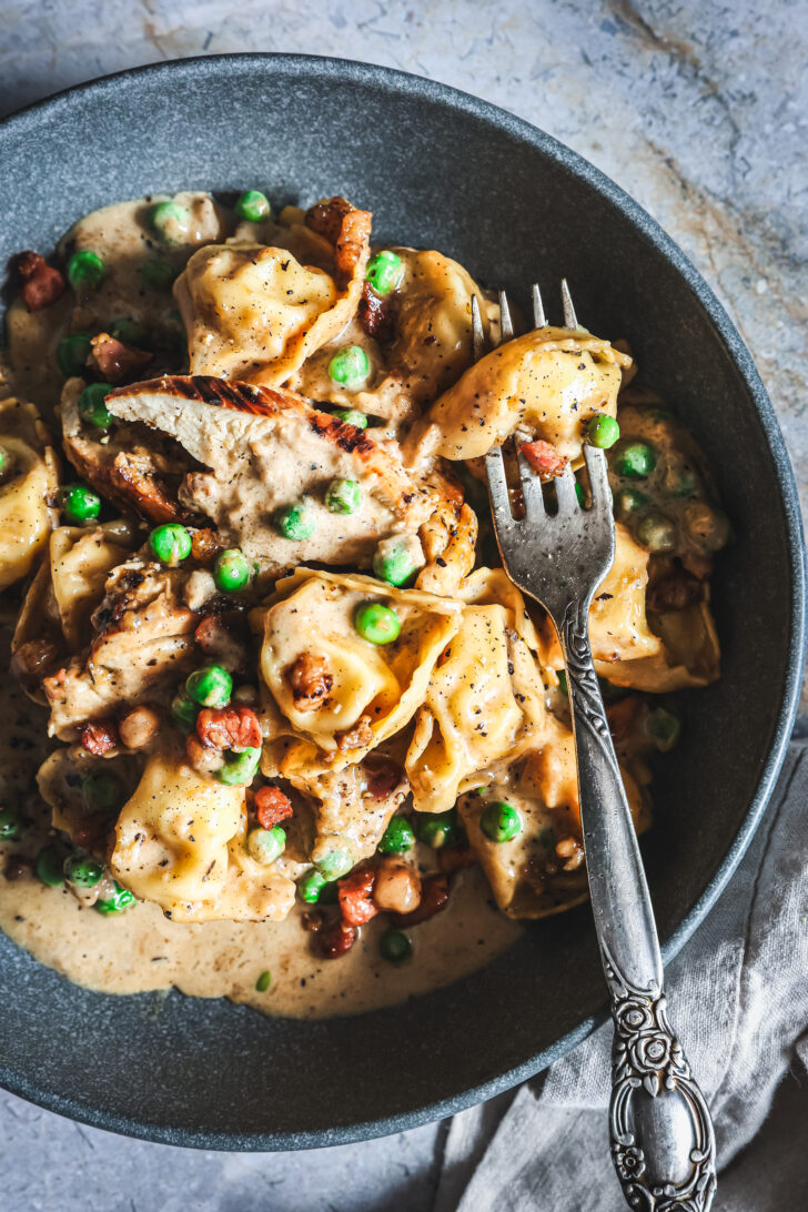 Close-up of chicken tortellini in creamy Alfredo sauce with peas and pancetta, served in a bowl with a fork.