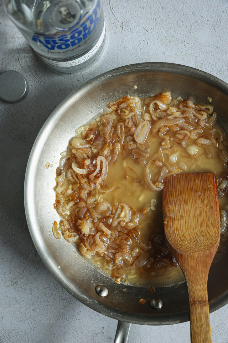 Caramelized shallots deglazed with vodka in a skillet, with a wooden spatula and a vodka bottle in the background