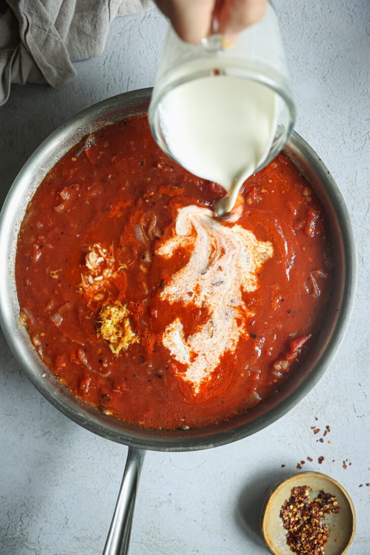 Heavy cream being poured into a skillet of simmering tomato vodka sauce, with chili flakes on the side for seasoning.