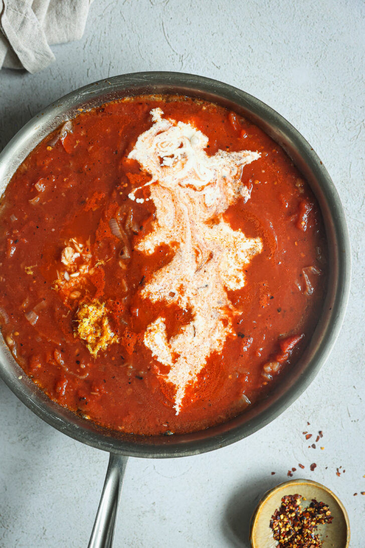 Rich tomato vodka sauce in a skillet, swirled with heavy cream and garnished with lemon zest and chili flakes on the side