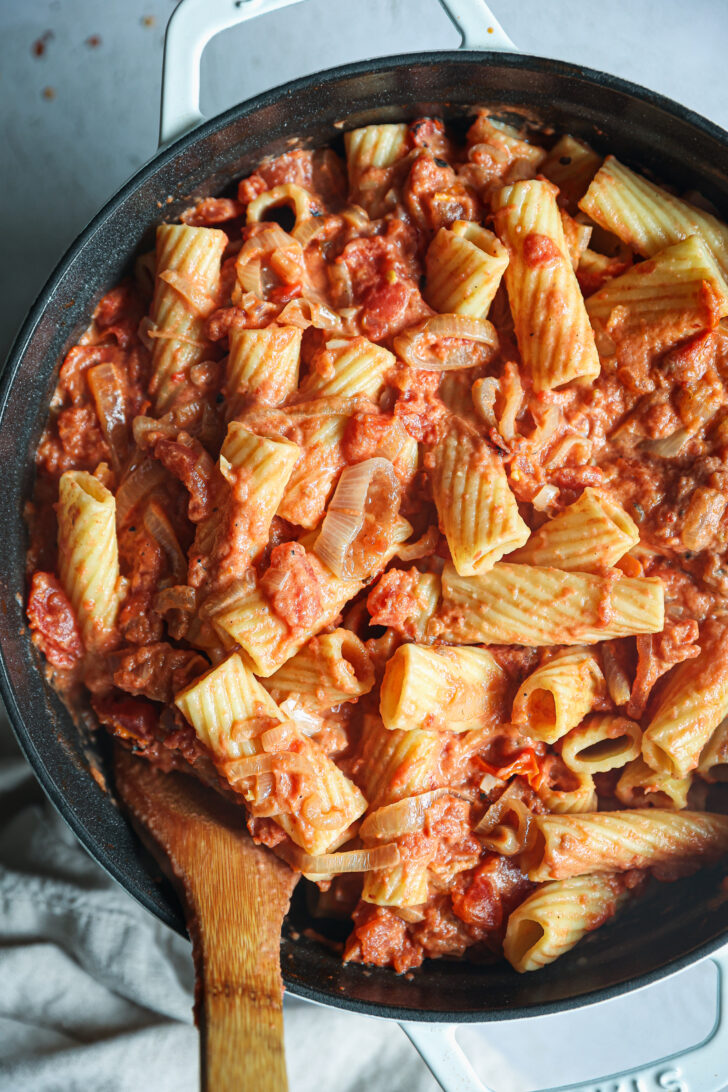 Rigatoni pasta tossed in creamy vodka sauce with caramelized shallots and tomatoes in a skillet, stirred with a wooden spatula