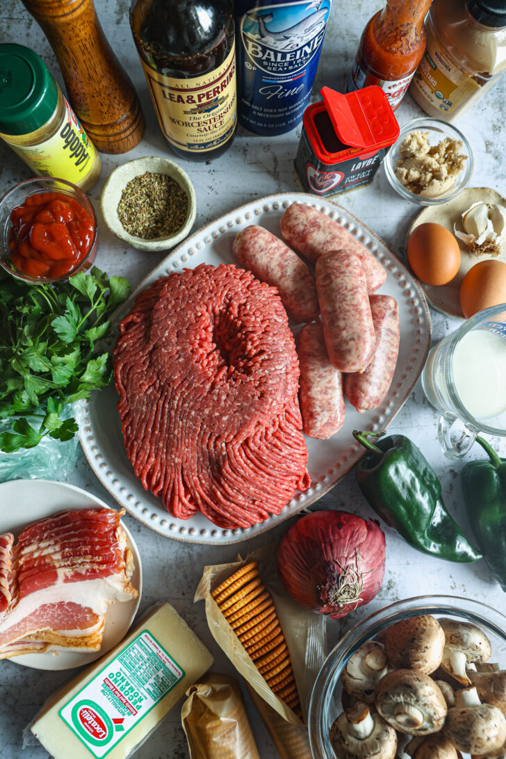 An overhead view of meatloaf ingredients, including ground beef, sausages, poblano peppers, herbs, spices, cheese, eggs, and crackers.