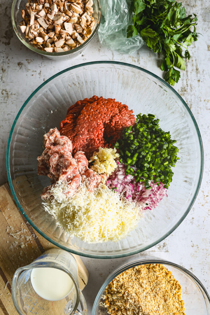 A large glass bowl with ground beef, sausage, diced peppers, onions, garlic, and shredded cheese, surrounded by breadcrumbs, milk, and parsley.