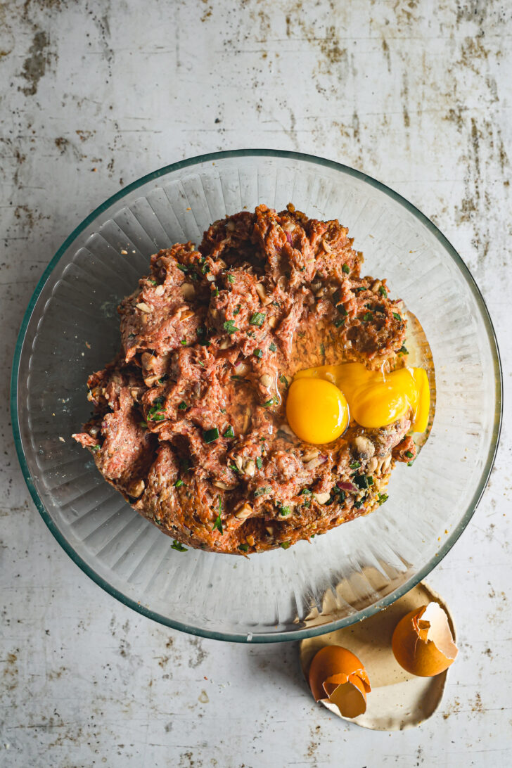 A glass bowl containing mixed meatloaf ingredients with two cracked eggs on top, next to broken eggshells on the counter.